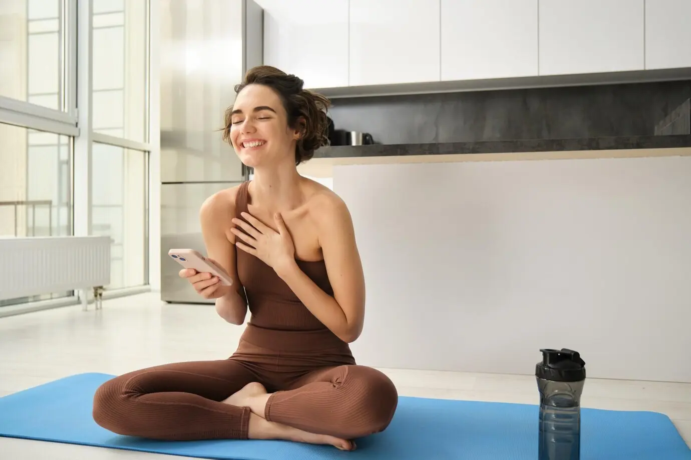 Bild einer sportlichen Frau in Sportkleidung, die zu Hause auf einer Yogamatte sitzt, ein Smartphone hält und eine Workout-App verwendet.