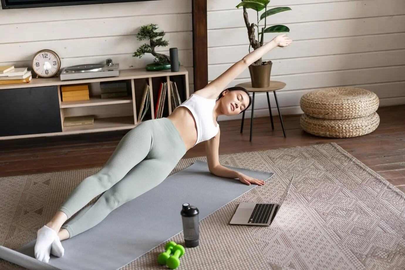 Eine junge asiatische Frau macht zu Hause auf einer Bodenmatte ein Workout und folgt dabei einem Sporttrainer in einem Video-Stream auf dem Laptop.