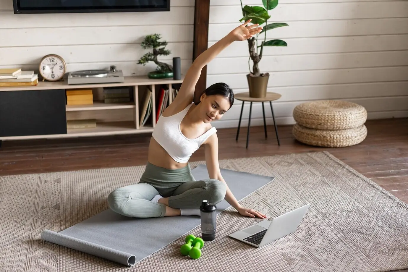 Eine junge asiatische Frau macht zu Hause ein Fitness-Workout mit Dehnübungen, streckt die Arme und schaut auf den Laptop.
